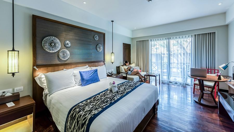 A modern hotel room with a bed, a large decorative headboard, and a seating area by the window.