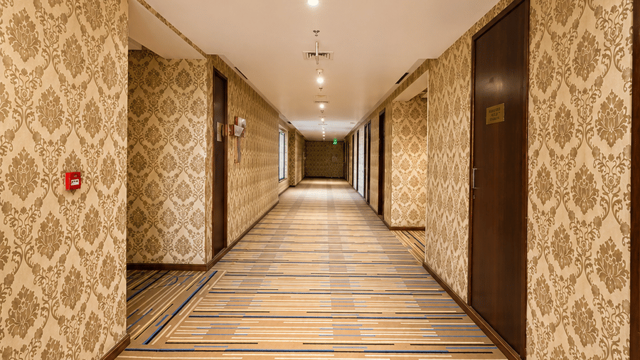 Long hotel corridor at Golden Tulip Kukas Jaipur with patterned carpet flooring, recessed ceiling lights, and decorative wallpaper lining both sides.