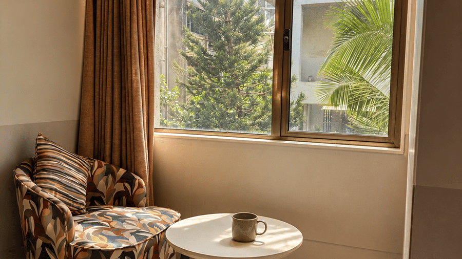 A seating space with a cushion, a table, and a window partially covered with curtains. | The Oriental Residency