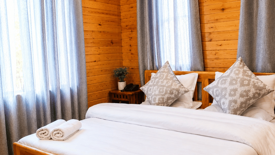 A Standard Room at Dawat e Drung by FrozenFall Resort, Drung, Tangmarg, featuring a wooden double bed frame, window curtains and a small geometric rug.