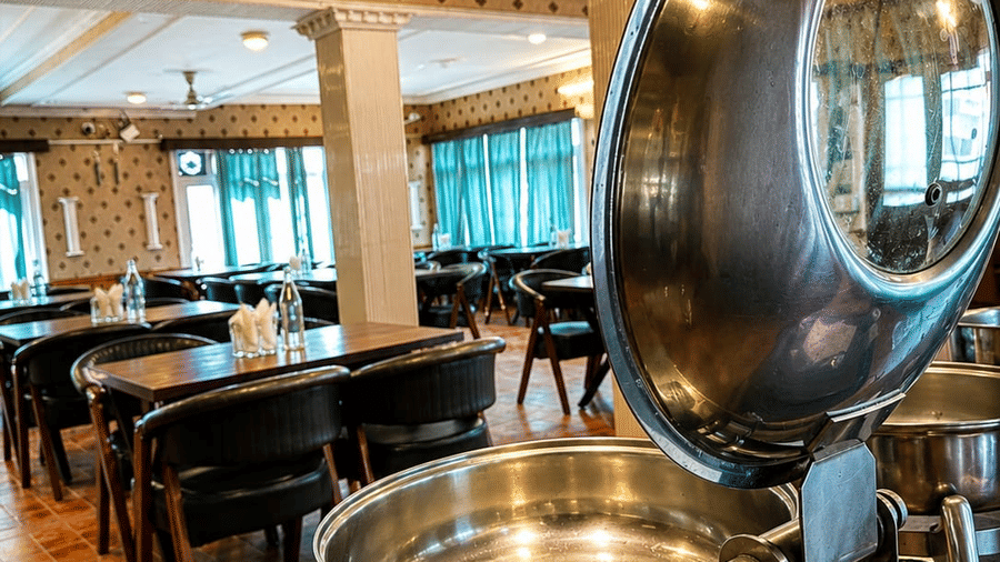 An indoor buffet area at Hotel Outlook, Pahalgam, showing a metal chafing dish with an open lid, set in front of dining tables and chairs.