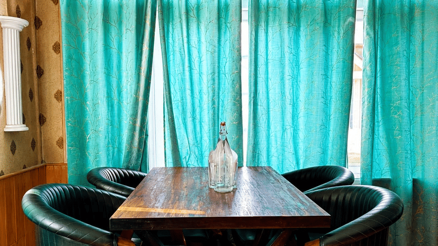 A dining setup at Hotel Outlook, Pahalgam, showing a wooden table surrounded by chairs positioned in front of windows with curtains.