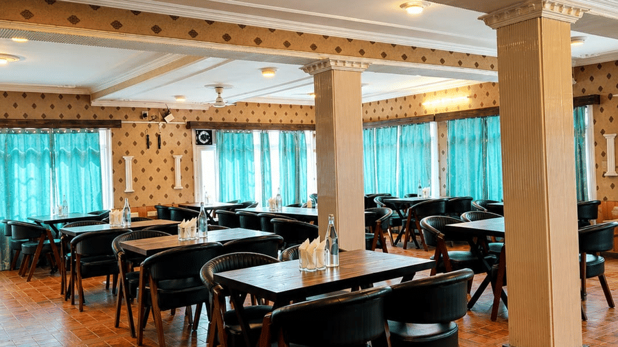 A dining area at Hotel Outlook, Pahalgam, with rows of tables and chairs arranged on a patterned floor under a ceiling with support pillars.