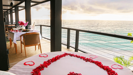 Romantic oceanfront bed adorned with rose petals arranged in a heart shape, set on a terrace overlooking the sea at sunset.