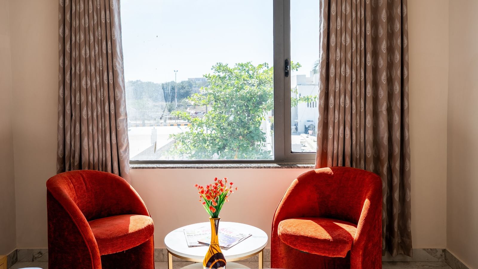 A cosy seating area at Daksh The Grand Gowardhan, Dwarka, with two vibrant red chairs by a window along with a coffee table, perfect for relaxation.