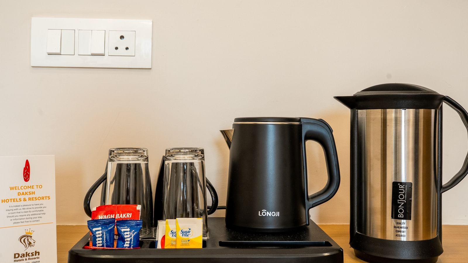 A tray kept on a table at Daksh The Grand Gowardhan, Dwarka, with a kettle, cups, and tea/coffee supplies with an electrical socket beside it.