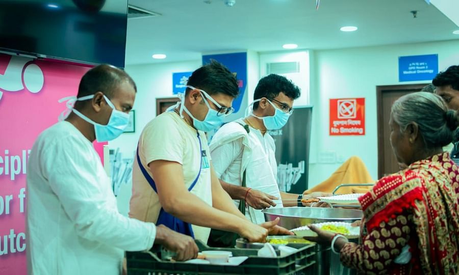 Volunteers wearing masks and aprons serve meals to a woman at HCG Hospital.