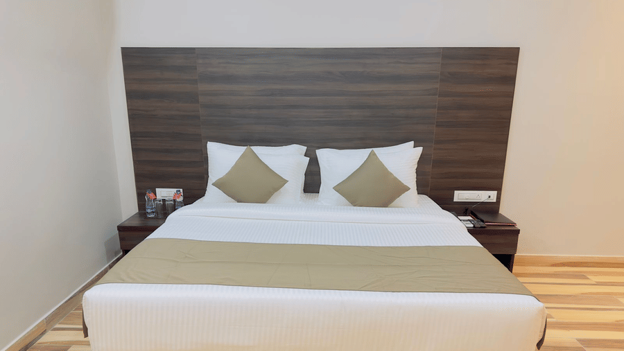 A double bed with white and beige linens and a dark wooden headboard is centred in a room - Kankidham Lords Inn