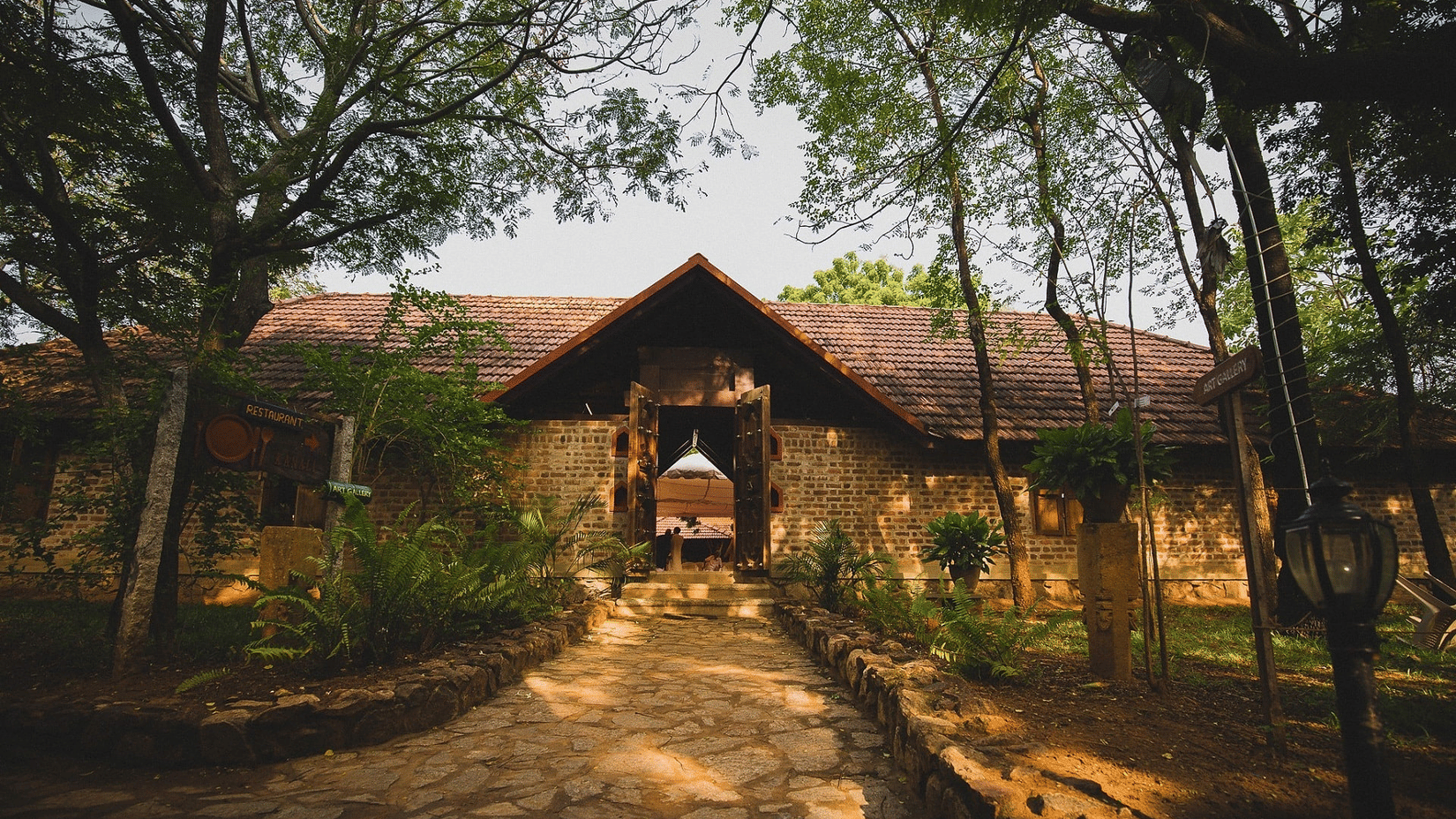 A stone path leads toward the entrance of a building with a pitched roof. Trees surround the structure and cast shadows on the ground.