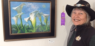 A woman in a black hat smiling next to her framed painting of white flowers, with a ribbon attached to the artwork.