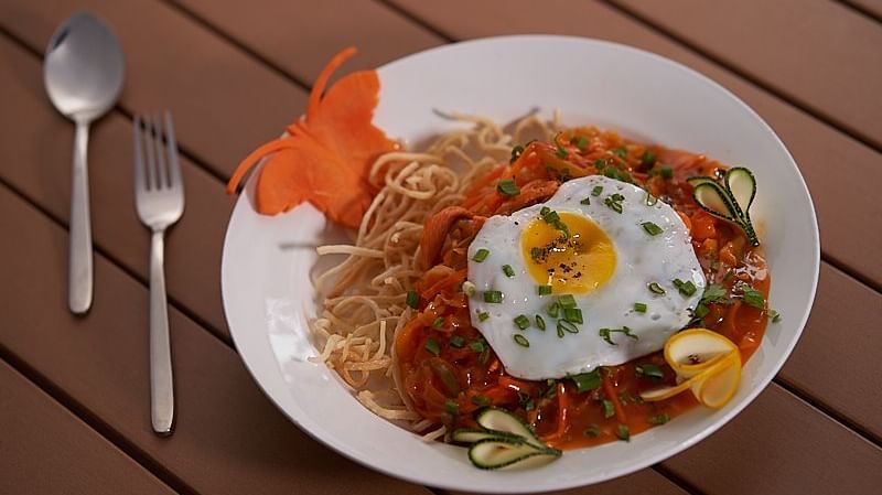  A plate of spaghetti topped with tomato sauce, a sunny-side-up egg, sliced vegetables, and half a boiled egg, served with a fork and spoon on a wooden table at The Hamuse Hotel, Kodaikanal.