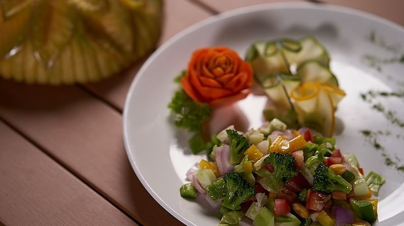 A plate of vegetable salad made with diced cucumbers, tomatoes, onions, and herbs, garnished with a carved tomato rose served on a wooden table at The Hamuse Hotel, Kodaikanal.