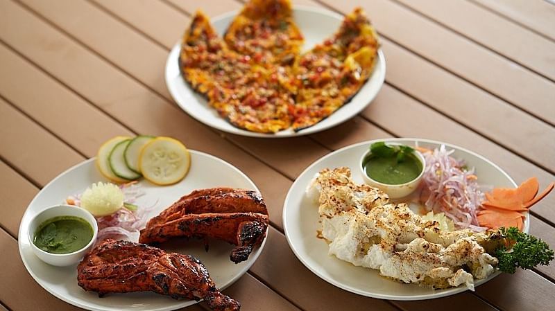 Three plates of food on a wooden table: a sliced vegetable pizza, grilled tandoori chicken with lime and green chutney, and a plate of crispy fried dish with sliced onions and green chutney at The Hamuse Hotel, Kodaikanal.