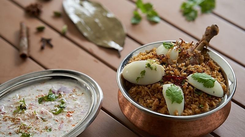 A copper bowl of biryani garnished with boiled egg slices and herbs, accompanied by a bowl of raita at The Hamuse Hotel, Kodaikanal.