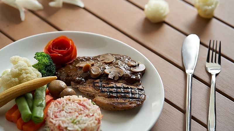 A plate featuring grilled meat, rice salad, mashed potatoes, steamed vegetables, and a tomato rose garnish, with cutlery placed beside it on a wooden table at The Hamuse Hotel, Kodaikanal.