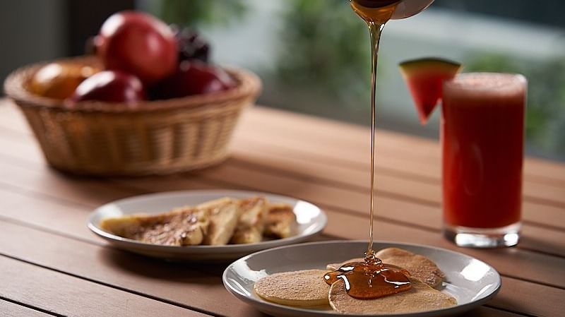 Honey dripping over a plate of pancakes on a wooden table with watermelon juice by side, and a bowl of fruits at The Hamuse Hotel, Kodaikanal.