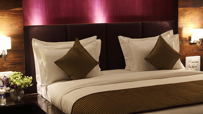 A warmly lit hotel room showcases a king size bed with a prominent dark headboard, crisp white linens, and decorative throw pillows.