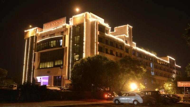 Night exterior of The Bristol Hotel, Gurgaon showing the multi-storey building outlined in string lights, the illuminated entrance, and cars parked in front.
