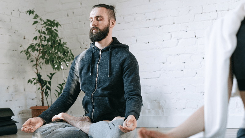 A person sitting cross-legged on a yoga mat with eyes closed, meditating in a calm indoor space with a plant and white brick wall in the background.