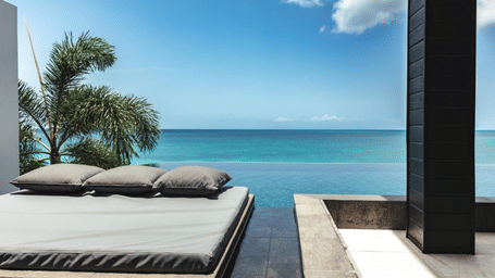 A modern outdoor bed on a patio overlooks a clear blue ocean under a bright sky.