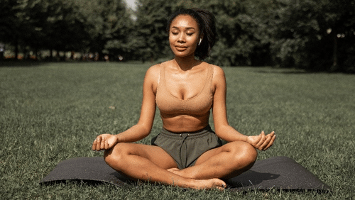 A woman meditates outdoors in a park, sitting cross-legged on a yoga mat with her eyes closed, smiling gently