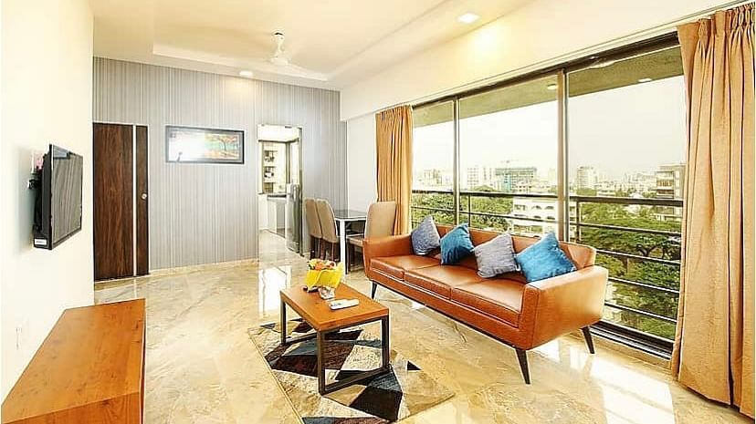 A well-lit living room with a leather sofa, blue cushions, a marble floor, a flat-screen TV on the wall and large windows that offer a view of the city.