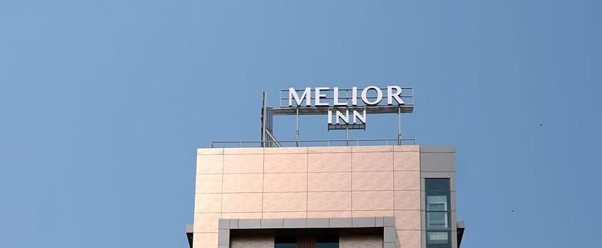 A tall, modern building against a clear blue sky, surrounded by some greenery - Melior Inn