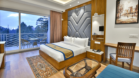 Image of the luxury room with balcony with a bed, bed side tables, chair, work desk, and a glass door leading to the balcony with a scenic view in the background - DLS Ark Holiday Inn, Dalhousie