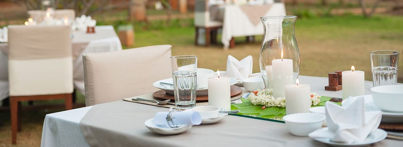 An outdoor dining setup with white tablecloths and chairs, green lawn and trees in the background - Shreyas Retreat