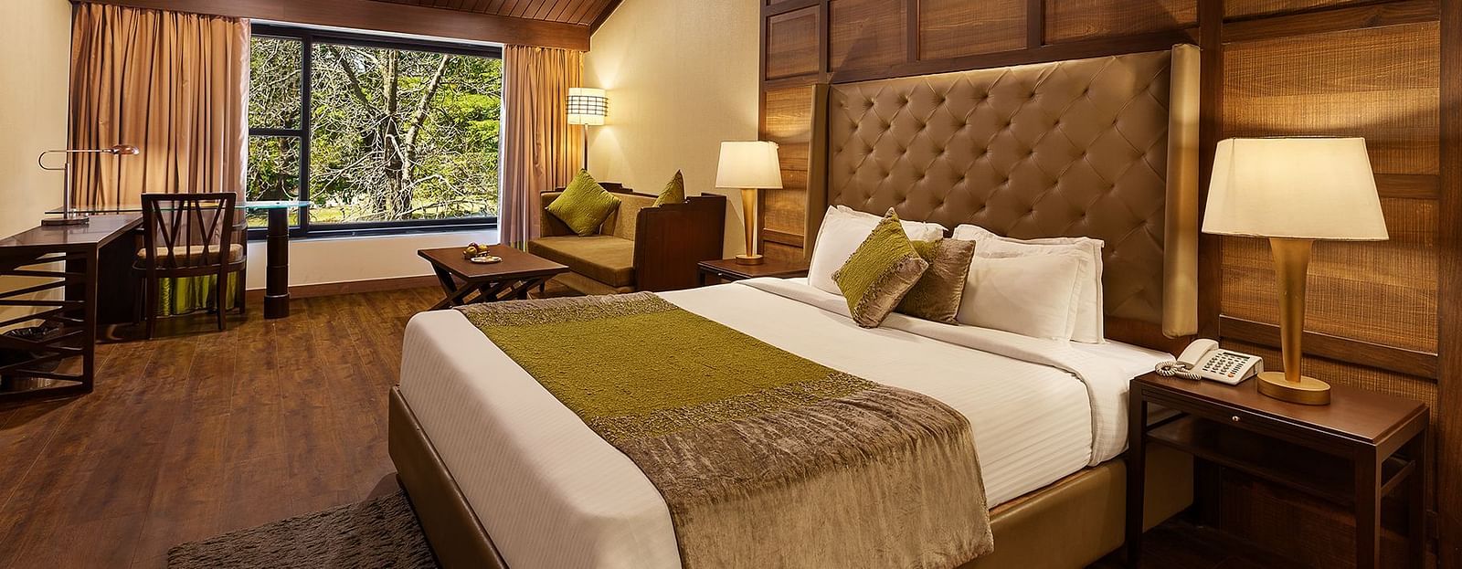 A bedroom with a large bed and a workdesk and chairs on the corner of the room, with large windows with trees visible - The Earl's Court, Nainital