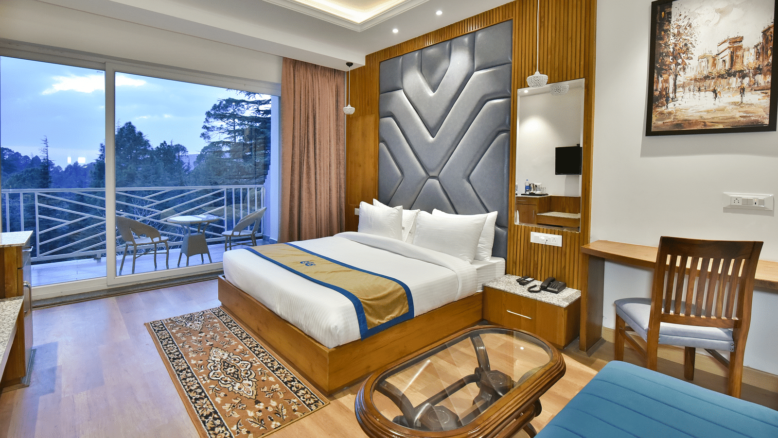 Image of the luxury room with balcony with a bed, bed side tables, chair, work desk, and a glass door leading to the balcony with a scenic view in the background - DLS Ark Holiday Inn, Dalhousie