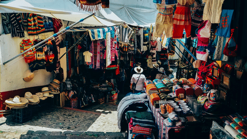 A street market with stalls selling clothes, bags, and textiles, with items displayed on tables and hanging from the stall frames.