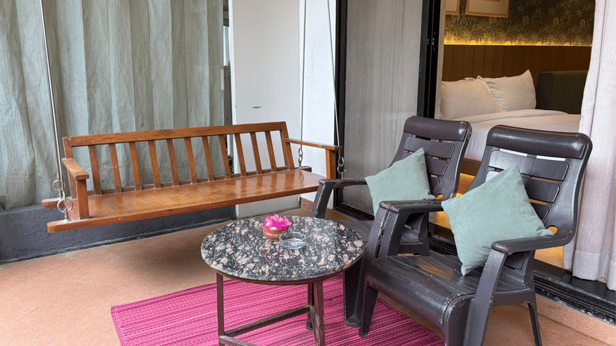 A wooden swing bench, 2 armchairs with cushions, and a small round table rest on a pink outdoor rug on a private terrace at Bougainvillea, Alibaug.