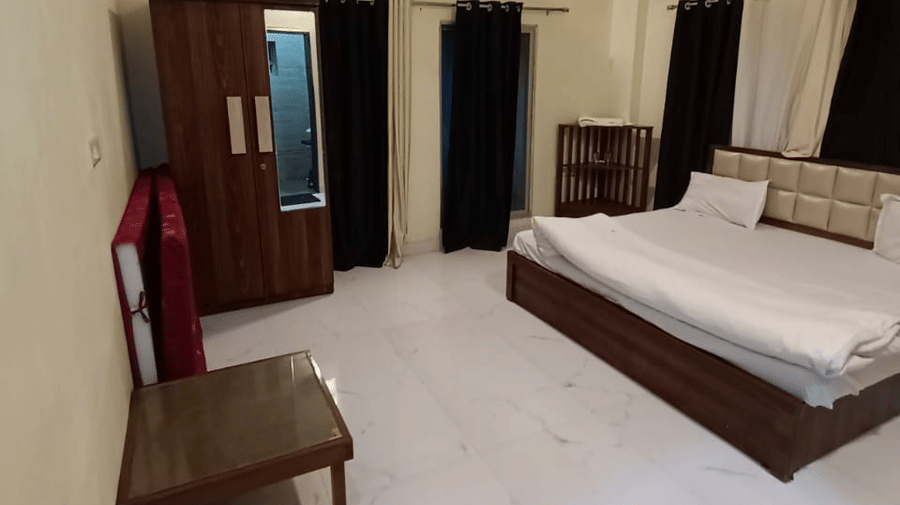A spacious bedroom with cream-coloured walls, a large bed with white sheets, dark wood furniture including a dresser and mirror, and a black door mat on the floor | Nandan Resort