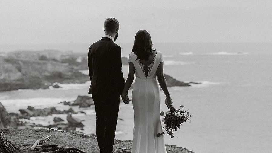 A black and white image of a couple gazing at the ocean from a rocky shoreline