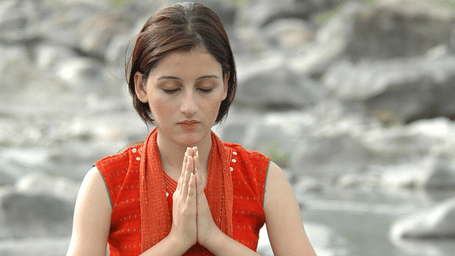 A woman sitting cross-legged on a large rock near a river with eyes closed and hands in prayer pose.