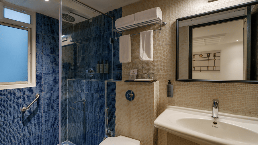 Modern bathroom at Shilton Suites, Bengaluru, featuring a glass enclosed walk in shower, wall mounted toilet, vanity with countertop basin and mirror.