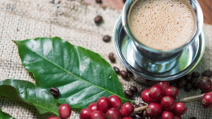 A steel cup of coffee alongside coffee beans and leaves on a piece of cloth