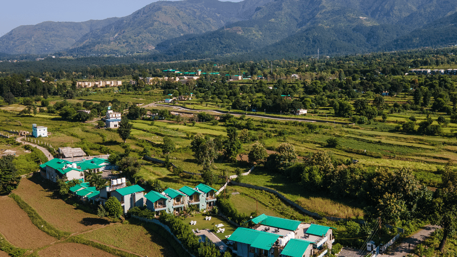 Aerial view of Corbett Nirvana Resort, Uttarakhand featuring cottage style buildings in clusters surrounded by farmlands, trees, and a backdrop of green hill.