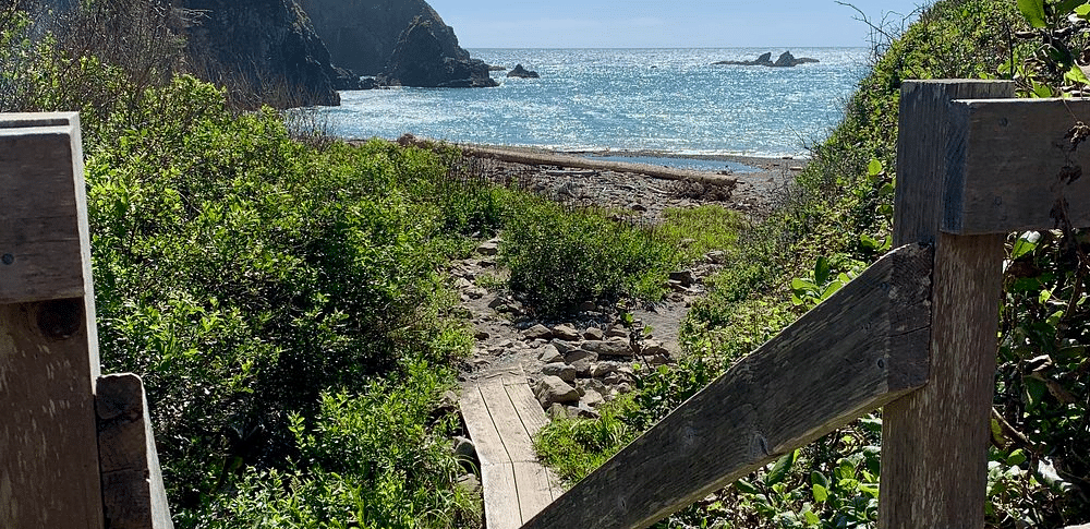 A path descends via steps towards a beach framed by cliffs and vegetation, under a partly cloudy sky.