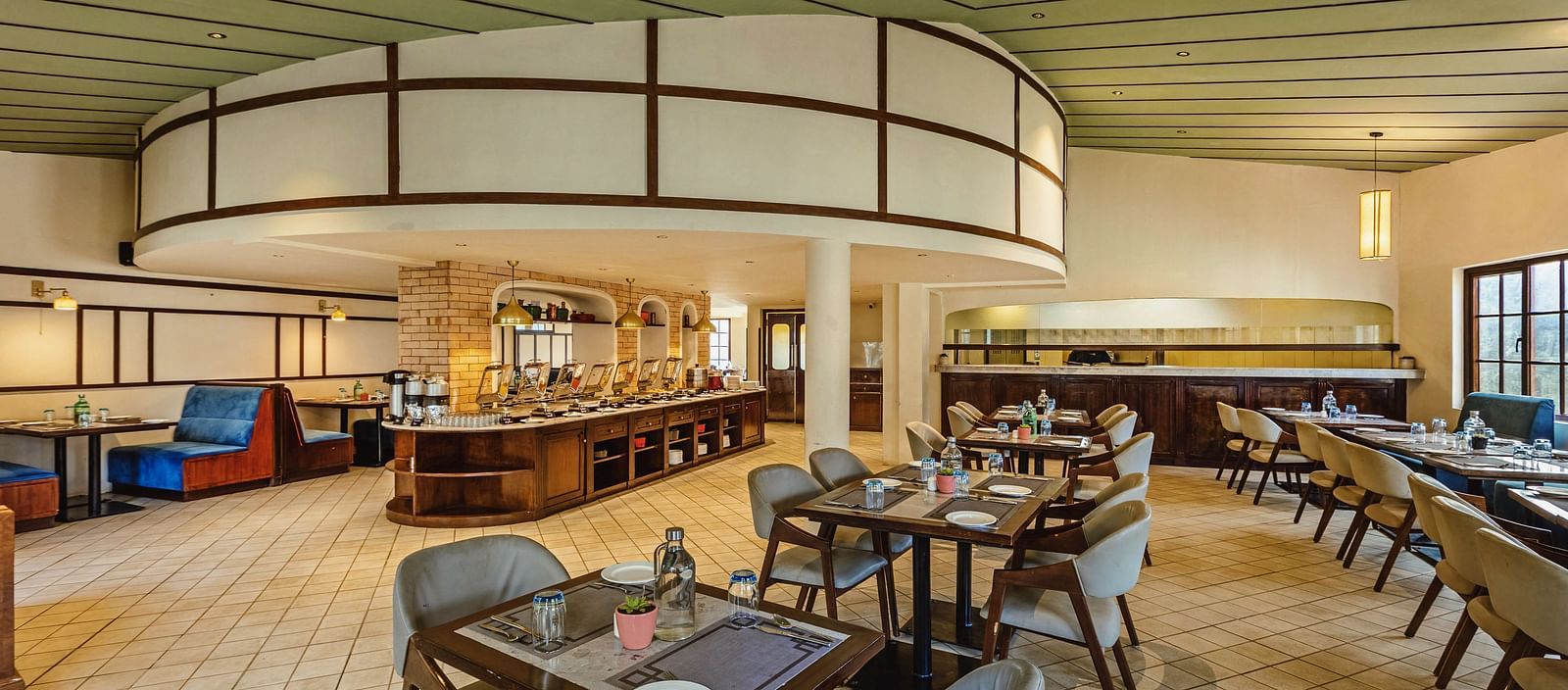 Spacious restaurant interior at Mango Hill Shola, Ooty, showcasing a curved buffet counter, comfortable booth seating, and wooden ceiling panels in a well-lit dining space.