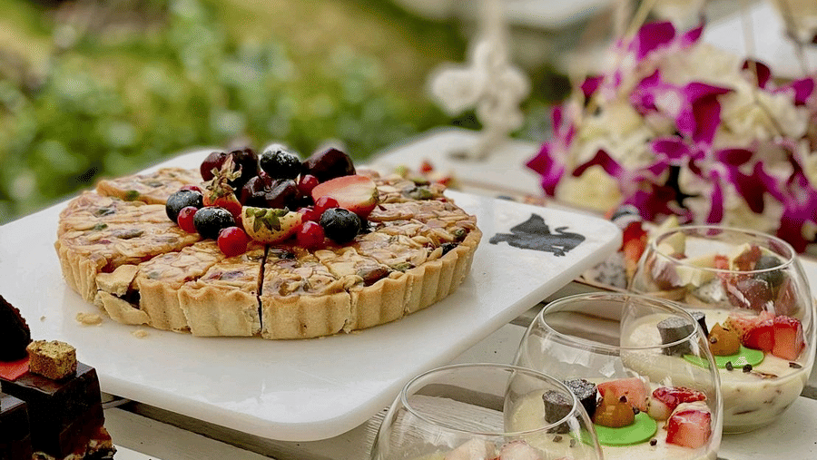Outdoor dessert spread featuring fruit tart, cream desserts, eclairs, and assorted pastries arranged on a garden table.