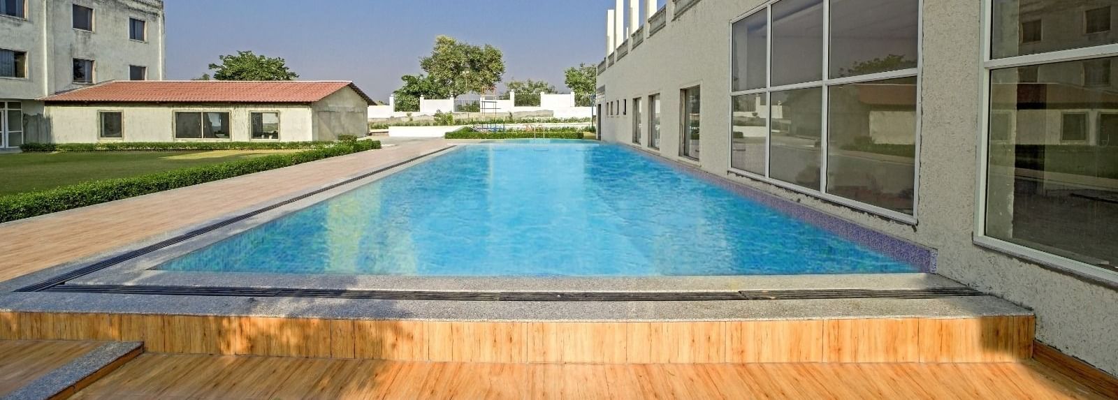 Outdoor swimming pool at Grassfield Riveria Resort, Jaipur, surrounded by modern buildings and a wooden deck area.