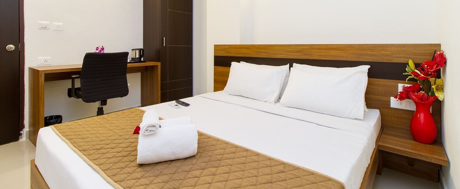 A hotel room including a double bed with brown bedding, a dark wardrobe, and a work desk and chair at Sanctum Suites Whitefield, Bangalore.