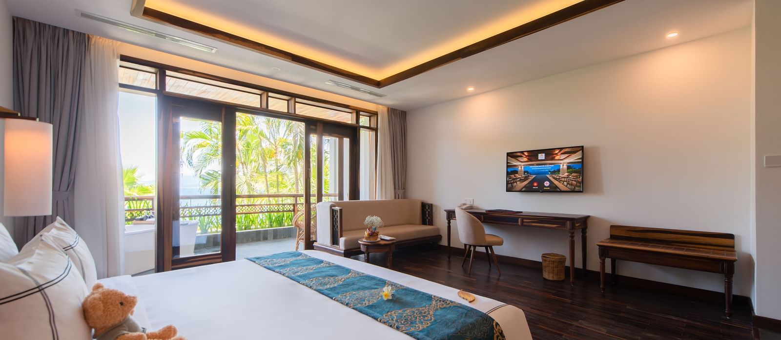 A hotel room with a large bed facing a television, furniture to relax and a balcony with a view of the greenery - Alibu Resort Nha Trang