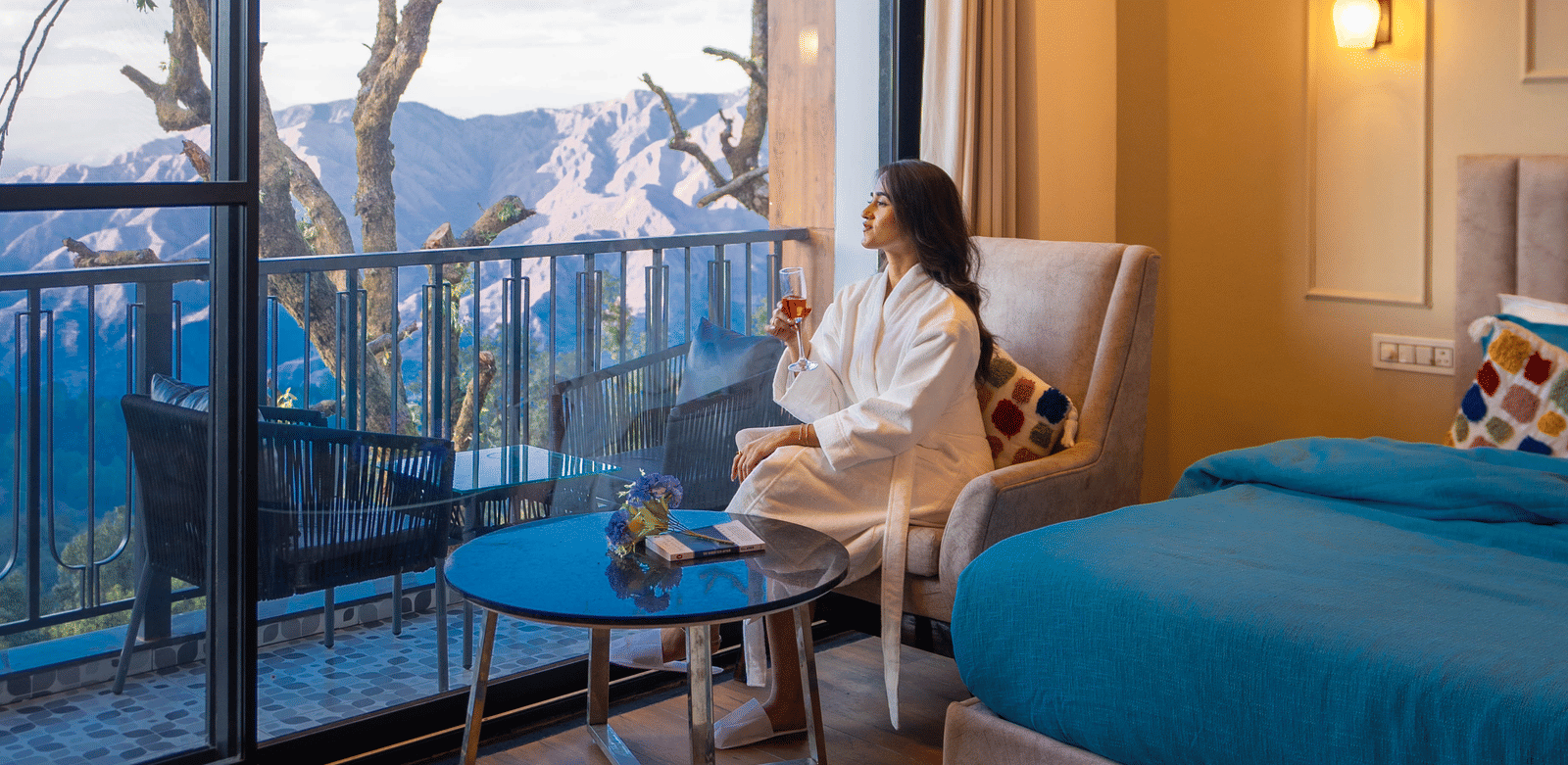 A woman dressed in a bathrobe looking outside of the Executive Room With Balcony & Valley View as she is sitting on an armchair at Perfectstayz Premium Westend Mall Road, Mussoorie.