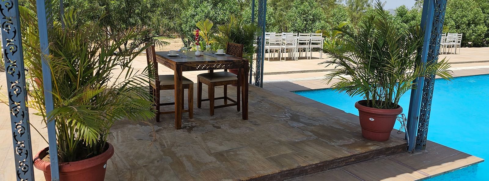 An outdoor table and two chairs on a raised platform next to a bright blue swimming pool - Bamboo Saa Mulberry Resort, Pushkar