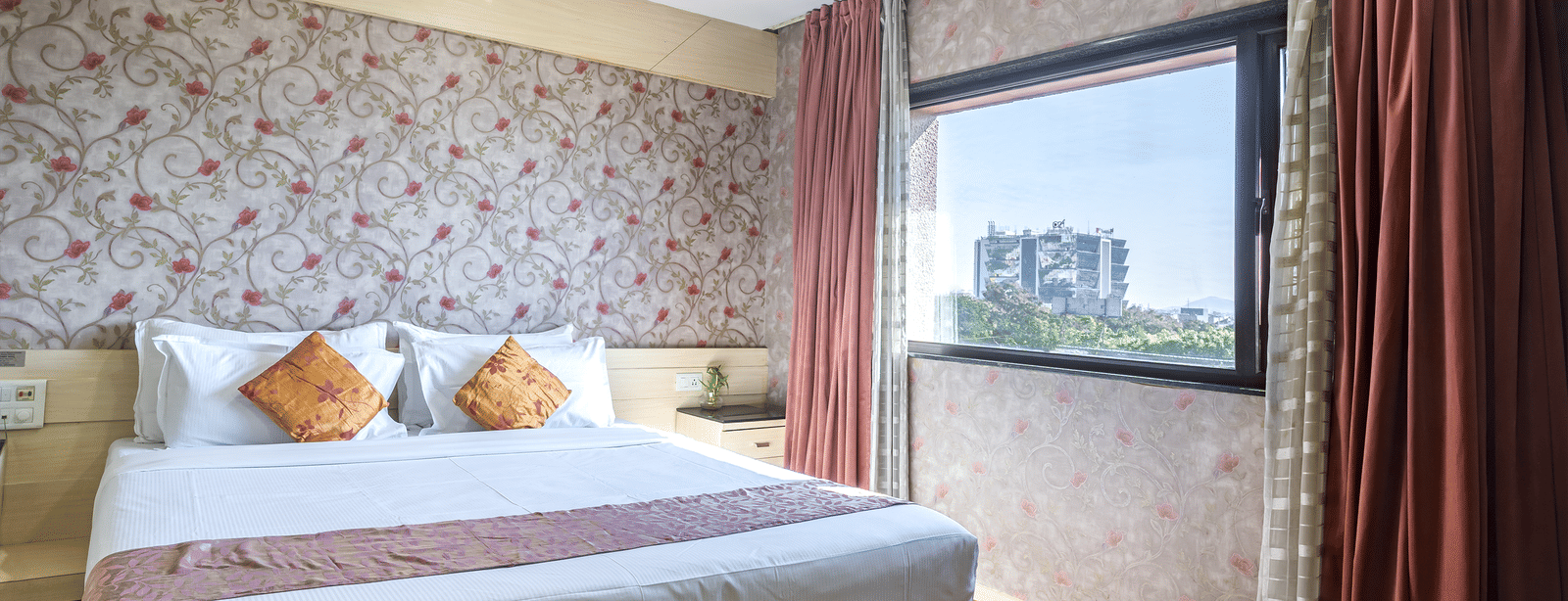 A cosy room with floral wall, king size bed, decorative cushions, a large window with maroon curtains, and light wooden flooring at Infiniti Hotel, Indore.