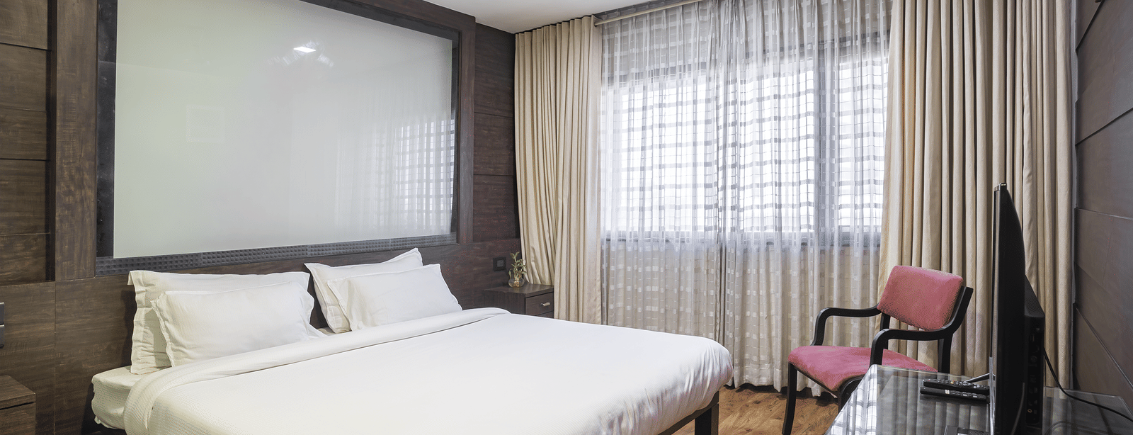 A spacious room with a king size bed, beige curtains, dark wall panelling, a pink armchair, and a T.V. at Infiniti Hotel, Indore.