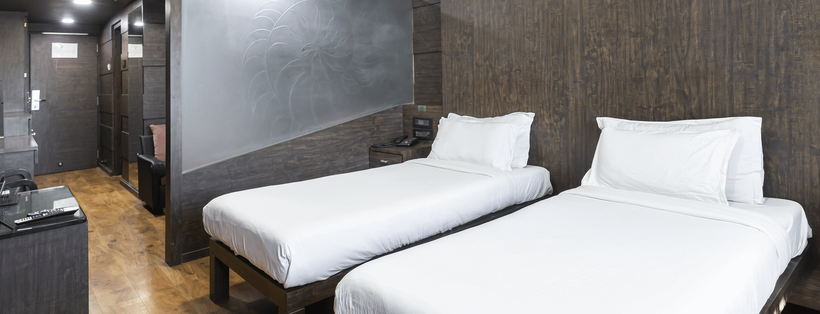 A room with two single beds, dark textured walls, a ceiling light, and a compact open wardrobe unit at Infiniti Hotel, Indore.